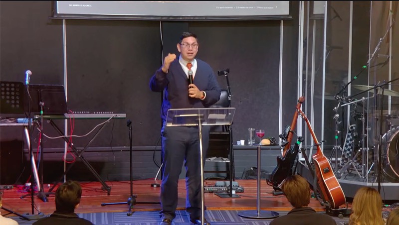 Predicador compartiendo el mensaje desde el púlpito durante el culto dominical, con instrumentos de alabanza y la mesa de la Cena del Señor en el escenario.