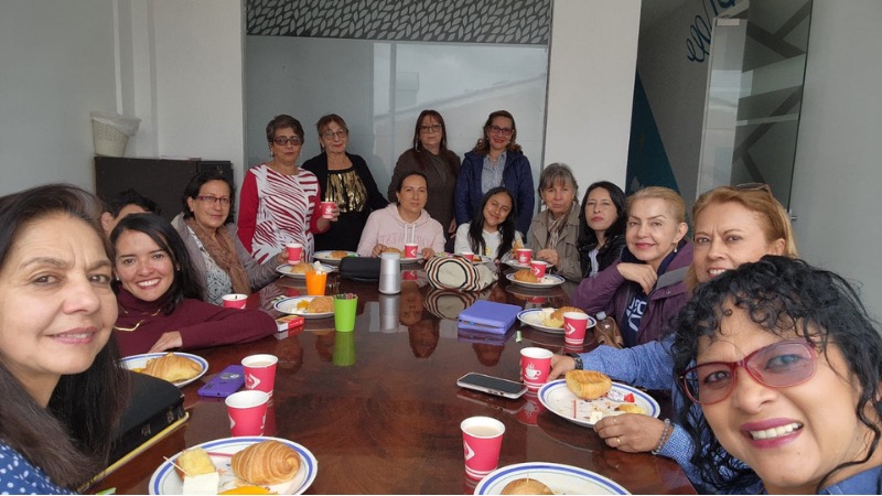 Mujeres de la iglesia sonriendo alrededor de una mesa compartida con café y pan durante una reunión de Siervas para su gloria.