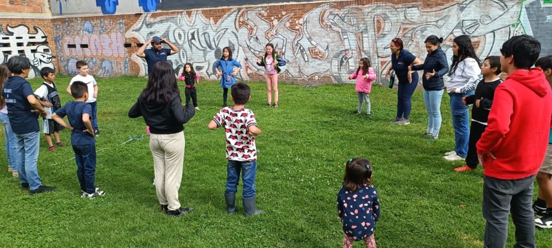 Niños y maestras jugando en círculo sobre el césped durante una actividad al aire libre de Oidores y hacedores.